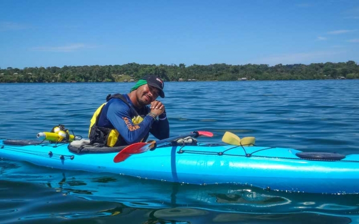 A person takes a break from kayaking on blue water to smile at the camera. In the background, there is a tree-lined shore.